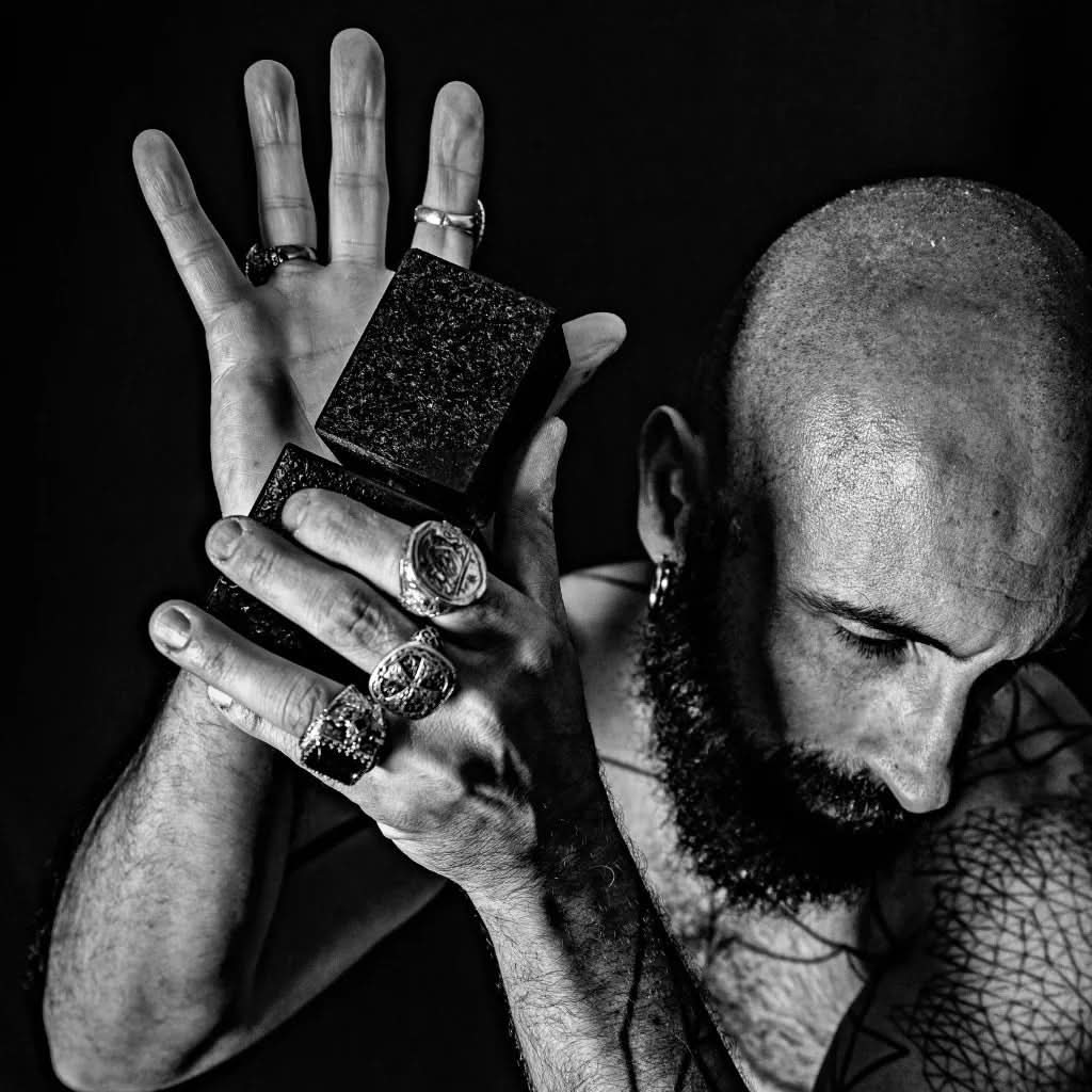 Czarno-białe zdjęcie mężczyzny z brodą i pierścionkami, trzymającego flakon perfum niszowych.