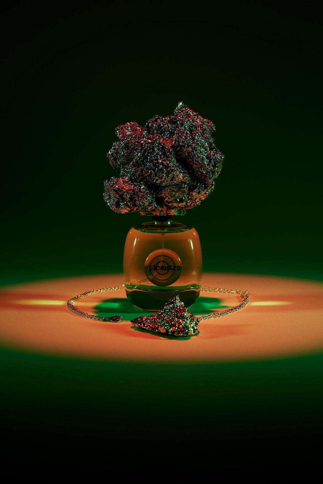 Butelka perfum niszowych Epicentro z kolorowym kamieniem i łańcuszkiem na tle zielono-pomarańczowym